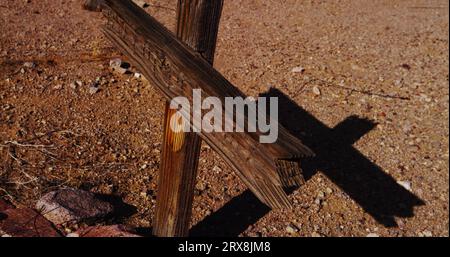 L'ombre rampante d'une croix tombale en bois traverse le trottoir naturel du désert dans un vieux cimetière ouest. Banque D'Images