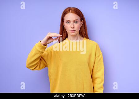 Belle femme rousse confiante sérieuse en chandail jaune montrant lettre D symbole de langue des signes pour l'humain sourd avec fond bleu. isolé Banque D'Images