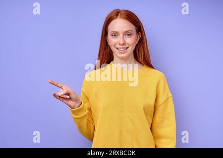 Belle femme rousse confiante heureuse en pull jaune élégant démontrant lettre Z symbole de langue des signes pour l'homme sourd avec fond bleu. iso Banque D'Images