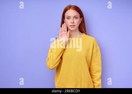 Jeune femme sourde muette aux cheveux rouges utilisant la langue des signes sur fond bleu, belle fille debout avec la main tendue montrant le signe d'arrêt, empêchant Banque D'Images