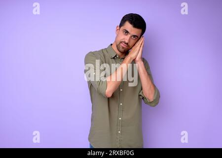 bel homme sérieux montrant geste de sommeil, couché sur les mains pliées regardant la caméra. guy faisant signe endormi rire portrait closeup isolé bleu Banque D'Images