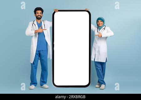 Publicité médicale Médecins arabes souriants en uniforme pointant vers le smartphone Big Blank Banque D'Images