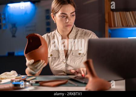 Femme archéologue faisant de la recherche travaillant tard la nuit dans le bureau, utilisant un ordinateur portable, analysant des artefacts de culture de civilisation antique Banque D'Images