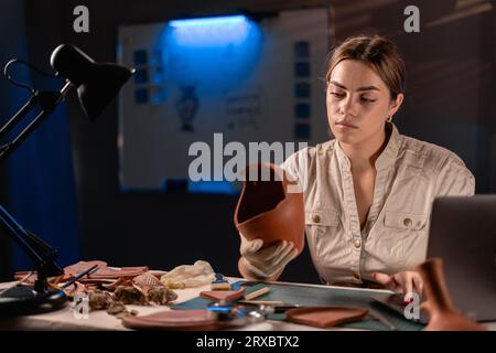 Femme archéologue faisant de la recherche travaillant tard la nuit dans le bureau, utilisant un ordinateur portable, analysant des artefacts de culture de civilisation antique Banque D'Images