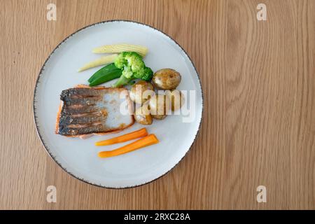 Une assiette de saumon poêlé accompagnée de pommes de terre nouvelles, de carottes, de brocoli et de maïs doux bébé est un choix nutritif. Le plat est présenté sur un morceau de bois. Banque D'Images