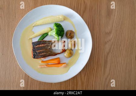 Une assiette de saumon poêlé servi avec des pommes de terre nouvelles, des carottes, du brocoli, du maïs doux et de la sauce au vin blanc est placée sur une table en bois. Banque D'Images