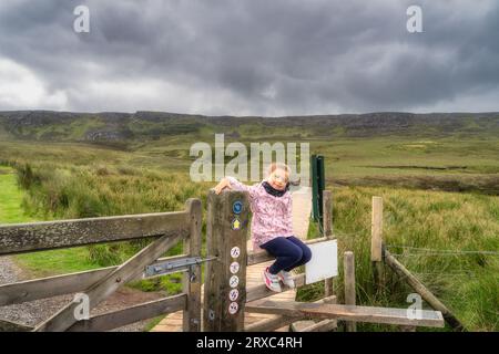 Jeune fille assise sur une porte en bois à Cuilcagh Boardwalk et Mountain Park avec des montagnes pittoresques en arrière-plan, Fermanagh, Irlande du Nord Banque D'Images