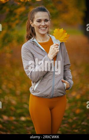 Bonjour automne. Portrait de femme moderne souriante dans des vêtements de fitness dans le parc avec feuille d'automne. Banque D'Images