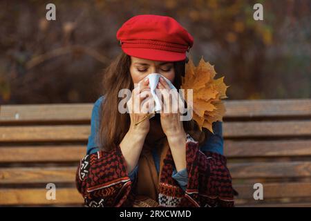 Bonjour automne. femme élégante en chapeau rouge avec feuilles d'automne et serviette soufflant nez tout en étant assise sur le banc dans le parc de la ville. Banque D'Images