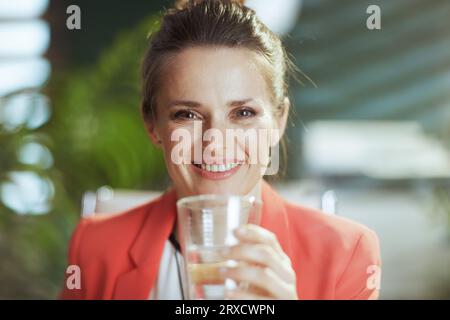 Lieu de travail durable. Portrait de femme d'affaires moderne heureuse au travail dans une veste rouge avec tasse d'eau. Banque D'Images