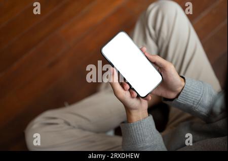 Image rapprochée d'un homme utilisant son smartphone tout en se détendant assis sur le sol dans une pièce. Maquette d'écran blanc de smartphone. Les gens et la technologie conce Banque D'Images