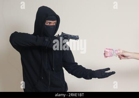 Portrait d'un homme mystérieux portant un sweat à capuche noir et un masque faisant des tirs avec une arme à feu tout en extorquant un billet de banque d'argent rupiah de la victime. Image isolée sur gr Banque D'Images