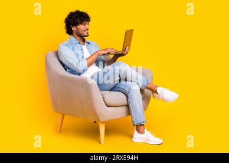 Photo pleine grandeur de l'homme intelligent habillé chemise en denim assis dans le fauteuil regarder à l'ordinateur portable faire la commande en ligne isolé sur fond de couleur jaune Banque D'Images