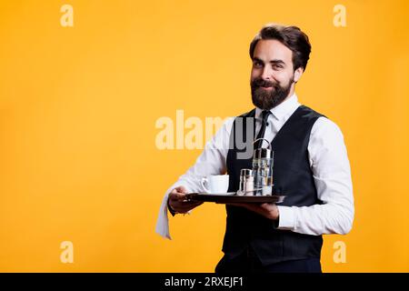 Élégant homme majordome tenant le plateau pour mettre le sel et le poivre sur la table au restaurant, portant la tasse de café sur le plateau pour les clients dans le studio. Serveur de luxe professionnel posant sur la caméra. Banque D'Images