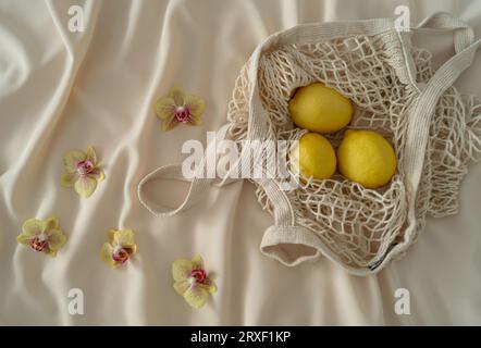 Poser à plat des orchidées tropicales et des citrons dans un sac de filet au crochet. Idée esthétique estivale. Concept créatif minimal. Banque D'Images