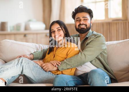 Heureux amoureux indiens profitant du temps ensemble à la maison, assis sur le canapé, embrassant et souriant à la caméra, espace libre Banque D'Images