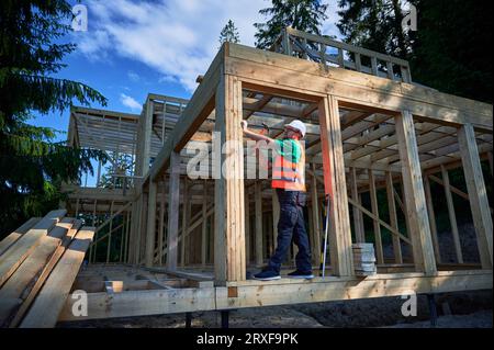 Menuisier construisant une maison à ossature en bois de deux étages près de la forêt. Homme martelant des clous dans la structure tout en portant un casque de protection et un gilet de construction. Concept de construction écologique moderne Banque D'Images