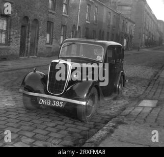 1960, historique, une voiture à moteur d'une époque antérieure, peut-être une Morris, garée dans une rue pavée de maisons victoriennes en terrasses à Shaw, Oldham, Manchester, Angleterre, Royaume-Uni. Remarquez les essuie-glaces simples en haut de la vitre côté conducteur et les portes arrière à charnières. On parle souvent de portes « suicide », car si la voiture avance et qu'elle s'ouvre, quiconque saisit la porte pour l'arrêter risque d'être éjecté hors du véhicule, car les voitures fabriquées à cette époque ne sont pas équipées de ceintures de sécurité. Banque D'Images