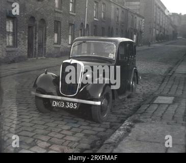 1960, historique, une voiture à moteur d'une époque antérieure, peut-être une Morris, garée dans une rue pavée de maisons victoriennes en terrasses à Shaw, Oldham, Manchester, Angleterre, Royaume-Uni. Remarquez les essuie-glaces simples en haut de la vitre côté conducteur et les portes arrière à charnières. On parle souvent de portes « suicide », car si la voiture avance et qu'elle s'ouvre, quiconque saisit la porte pour l'arrêter risque d'être éjecté hors du véhicule, car les voitures fabriquées à cette époque ne sont pas équipées de ceintures de sécurité. Banque D'Images