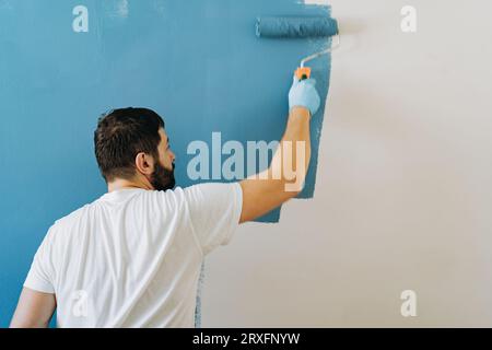 homme barbu caucasien peignant mur avec rouleau à peinture. Peinture appartement, rénovation de la maison avec peinture de couleur bleue. Image avec mise au point sélective Banque D'Images