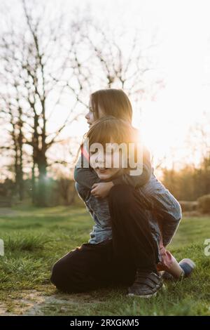 Charmant adolescent assis sur la pelouse verte dans le site rural tandis que la soeur embrassant frère de l'arrière et regardant loin. Garçon attentionné tenant charmante fille Banque D'Images