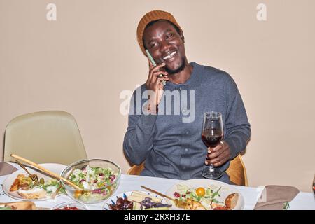 Portrait de jeune homme noir souriant parlant par smartphone à table à dîner, espace copie Banque D'Images