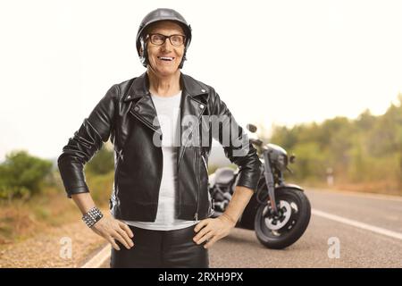 Vieux motard masculin avec un casque et une veste en cuir debout sur une route ouverte Banque D'Images