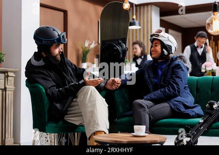 Petit ami et petite amie en possession d'équipement de sports d'hiver engagés dans le dialogue sur les plans pour le ski et le snowboard. Couple enthousiaste habillé chaleureusement pour les activités de neige assis dans le hall. Banque D'Images