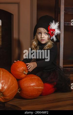 Une fille en costume de sorcière est assise près de la cheminée et joue avec des citrouilles. Halloween. Cadre vertical. Banque D'Images