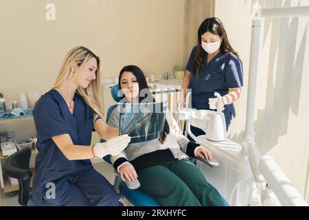 Latina femme dentiste expliquant Tooth X-Rays à Un patient, tandis que son assistante verse de l'eau pour le patient. Banque D'Images