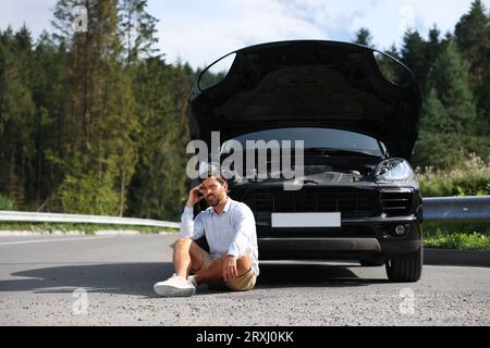 Homme stressé assis sur la route asphaltée près de la voiture cassée à l'extérieur Banque D'Images