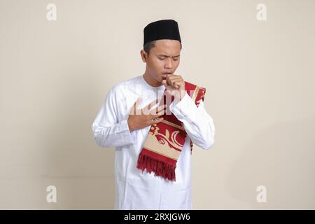 Portrait d'un homme musulman asiatique attrayant en chemise blanche gardant la paume de la main couvrant sa bouche tout en toussant avec l'autre main tenant sa poitrine. Isol Banque D'Images