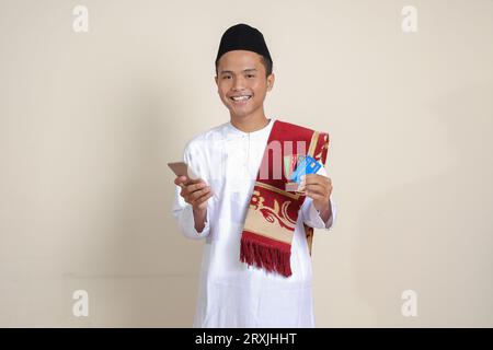 Portrait de l'homme musulman asiatique attrayant dans la chemise blanche avec skullcap tenant un téléphone portable et présentant la carte de crédit. Image isolée sur fond gris Banque D'Images