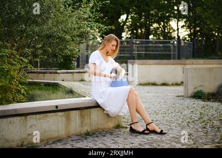 Femme blonde assise sur le banc et à la recherche de quelque chose dans son sac à main Banque D'Images