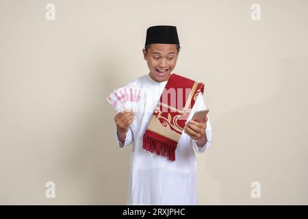 Portrait de l'homme musulman asiatique attrayant en chemise blanche montrant cent mille rupiah tout en utilisant un téléphone portable. Concept financier et d'épargne. Est Banque D'Images