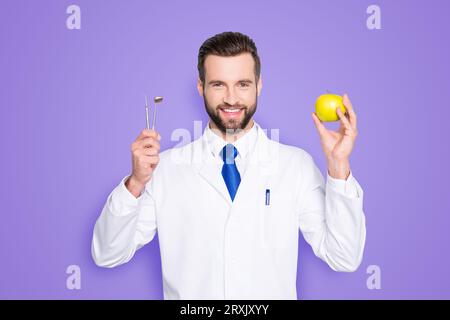 Portrait de dentiste toothy réussi en blouse de laboratoire blanche, cravate bleue, matériel de maintien et pomme verte dans les mains, isolé sur fond gris, recommander Banque D'Images