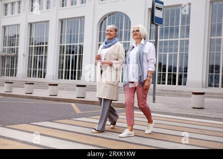 Deux femmes âgées aux cheveux gris élégamment habillées marchant, traversant la route en ville et souriant. Amis seniors à la mode et élégant à l'extérieur. Banque D'Images