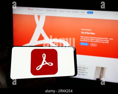 Personne tenant le téléphone mobile avec le logo du logiciel de gestion PDF Adobe Acrobat sur l'écran devant la page Web de l'entreprise. Concentrez-vous sur l'affichage du téléphone. Banque D'Images