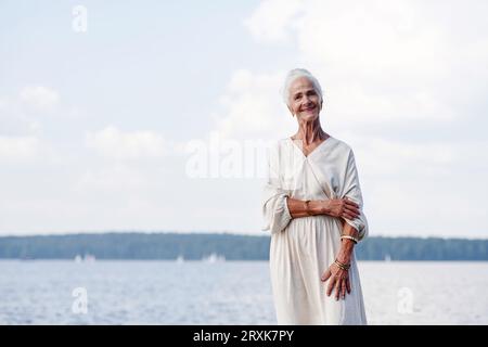 Portrait de femme senior élégante souriant à la caméra tout en se tenant dehors contre le lac Banque D'Images