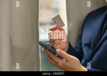 Photo recadrée de l'homme d'affaires tenant la carte de crédit et utilisant le téléphone portable, faisant des transactions en ligne ou des achats en ligne Banque D'Images