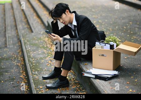 Employé masculin licencié avec boîte de biens à l'aide de smartphone sur les escaliers de l'immeuble de bureaux, perte de travail pendant la récession Banque D'Images