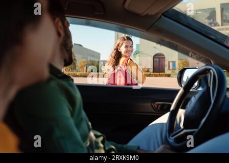 Mère et père déposant leur fille devant l'école, fille se retournant vers les parents assis dans la voiture et souriant Banque D'Images