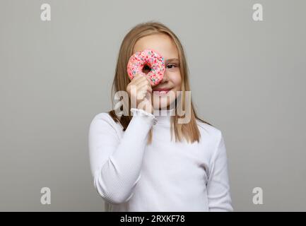 Portrait de jeune jolie fille joyeuse excitée en t-shirt blanc s'amusant avec des beignets sur les yeux sur fond gris. Enfance heureuse avec dessert savoureux. Banque D'Images