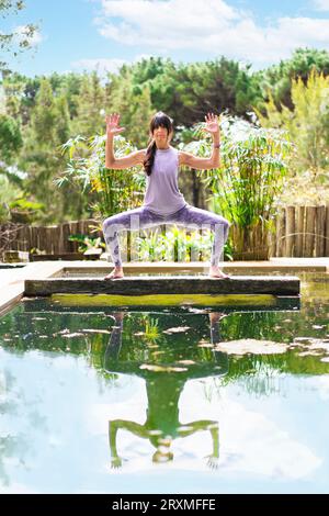 Une femme pratiquant le yoga pose Déesse avec Cactus Arms sur un tronc d'arbre au-dessus d'une piscine, tir vertical Banque D'Images