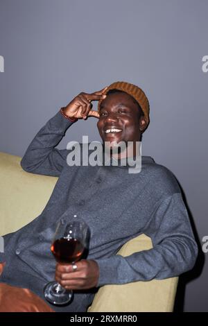 Portrait vertical de sourire jeune homme noir avec du vin rouge assis sur le canapé souriant à la caméra dans la boîte de nuit Banque D'Images