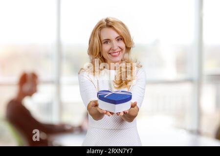 Heureuse femme blonde d'âge moyen souriante vous donne une boîte cadeau bleue. Bureau flou en arrière-plan. Banque D'Images