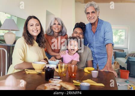 Une famille élargie joyeuse, comprenant des grands-parents, des parents et un enfant biracial, pose ensemble dans une cuisine spacieuse et ensoleillée pour un portrait de famille. Banque D'Images