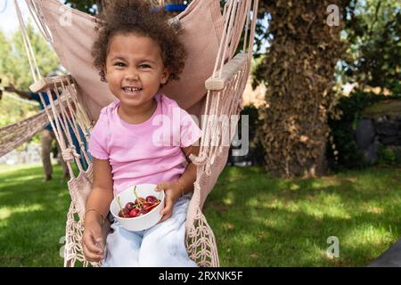 Une gaie fille de trois ans aux cheveux bouclés est assise sur une chaise à bascule en corde dans le jardin de la maison, tenant un bol de cerises. Elle est caucasienne et Afr Banque D'Images