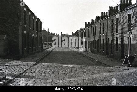 Dans les années 1960, les petites maisons en terrasses à façade plate bordent une rue pavée, les chalets de travailleurs pour les travailleurs dans les usines textiles locales, Oldham, Lancashire, Angleterre, Royaume-Uni. Banque D'Images