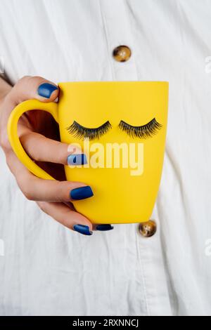 Salon de beauté. Femme méconnaissable avec manucure bleu vif tient dans sa main tasse jaune avec motif de cils pour l'extension. Logo, marque déposée. Soyez Banque D'Images